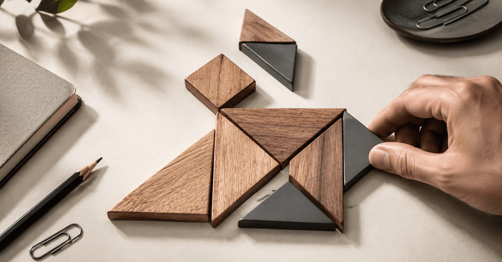 A wooden tangram on a desk being rearranged by hand into a new figure, the same pieces recomposed rather than removed